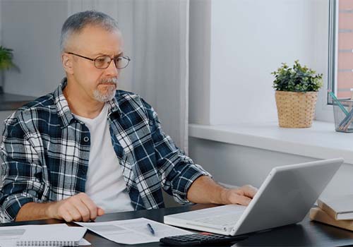Elderly Man Sitting At Laptop Looking At Bank Statements