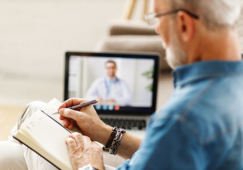 Elderly man writing in a notepad while making a video call from home to a mental health counselor.